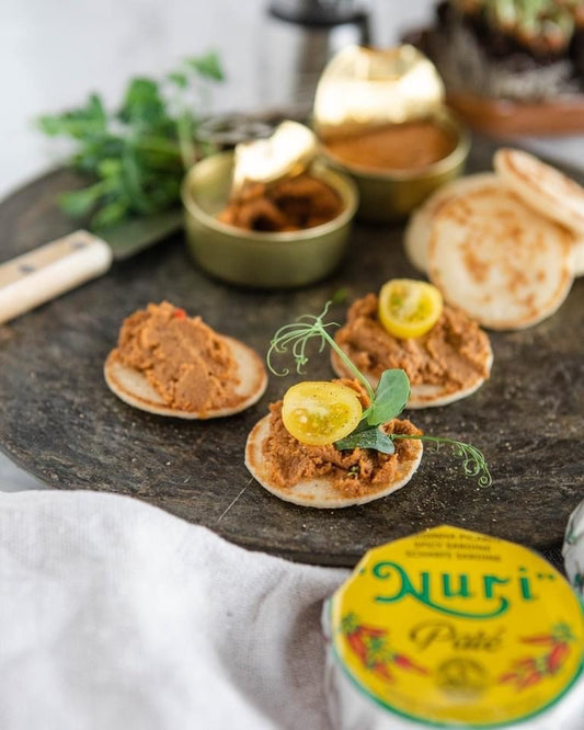 Kleine Pfannkuchen mit NURIs PIKANTE SARDINENPATÈ MIT OLIVENÖL, garniert mit Kräutern und einer gelben Tomatenscheibe auf einem dunklen, runden Tablett, die an die Aromen eines charmanten Fischrestaurants in Portugal erinnern.
