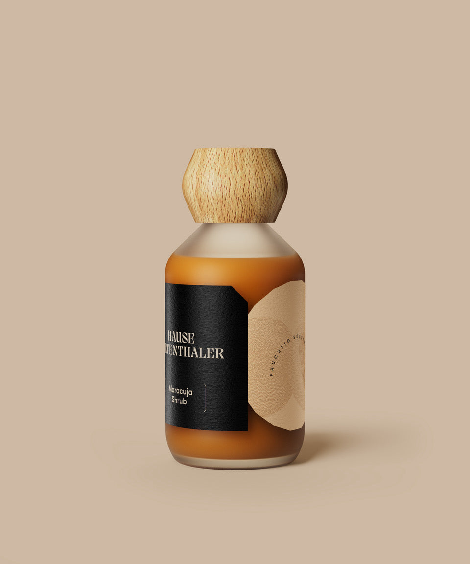 Eine braune Flasche mit Holzkappe und schwarzem Etikett, auf dem MARACUJA SHRUB von Haus Kaltenthaler abgebildet ist, vor einem beigen Hintergrund.