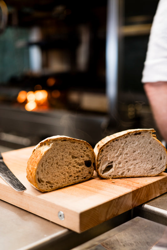 Zwei Hälften handwerklich hergestellten Brotes liegen auf einem MASSIVES KÜCHENBRETT AUS SCHWEDEN von Kockums Jernverk, in gemütlicher Küchenatmosphäre liegt ein Messer in der Nähe.