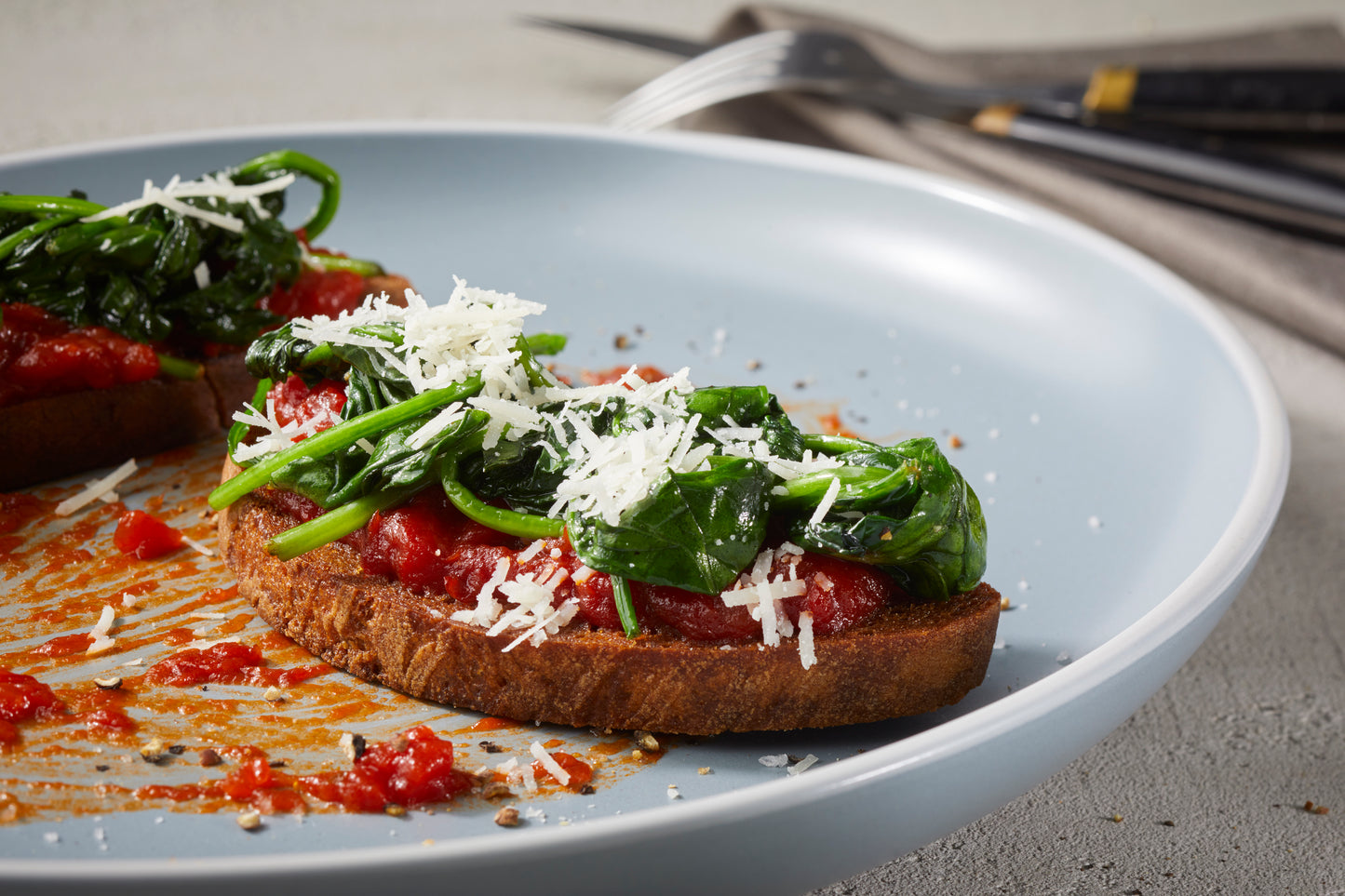 Zwei Scheiben Toast mit Rittmeyers Tomate Frito in Olivenöl, sautiertem Spinat und geriebenem Käse auf einem hellblauen Teller.