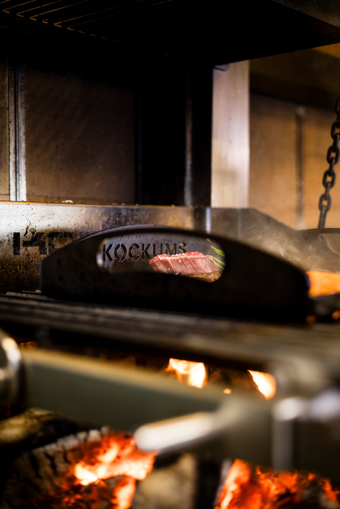 Ein Steak brutzelt auf einer Kockums Jernverk GRIDDLEPLATTE 35 x 35 cm, darunter flackern Flammen in der gemütlichen Küchenatmosphäre.
