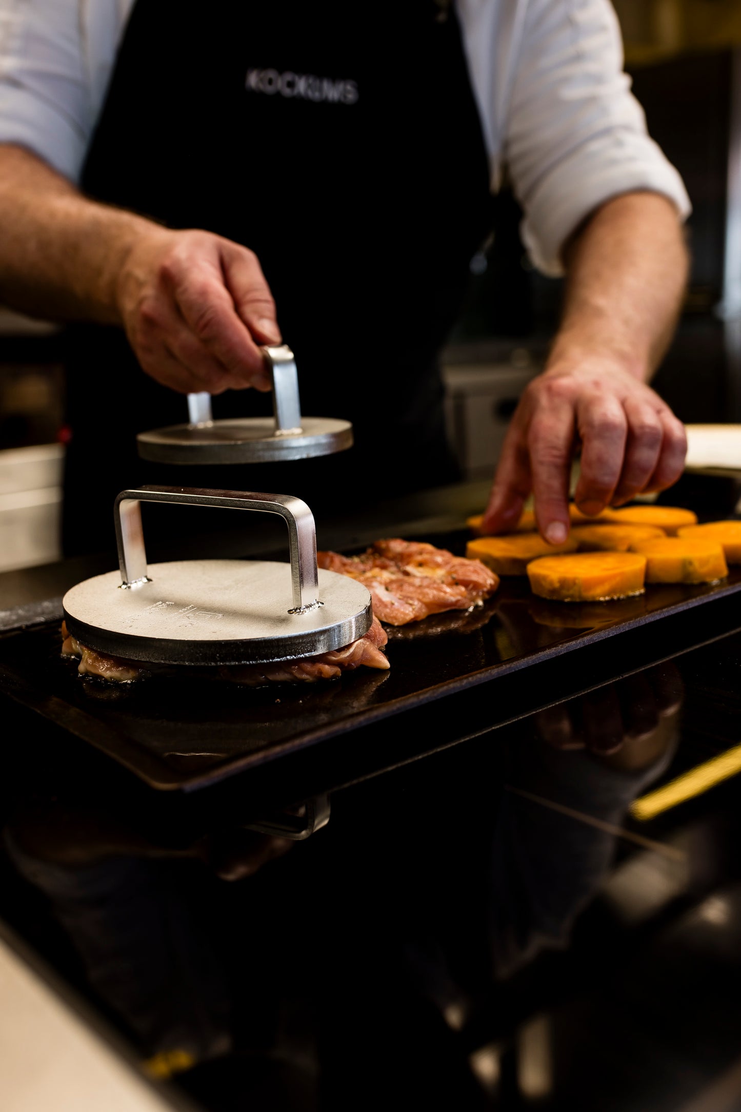 Der Küchenchef setzt das Kockums Jernverk EDELSTAHL-GRILLGEWICHT geschickt ein, um Burger zu pressen und Gemüse auf einer großen Grillplatte fachmännisch zu grillen, um perfekte knusprige Braten zu erzielen.
