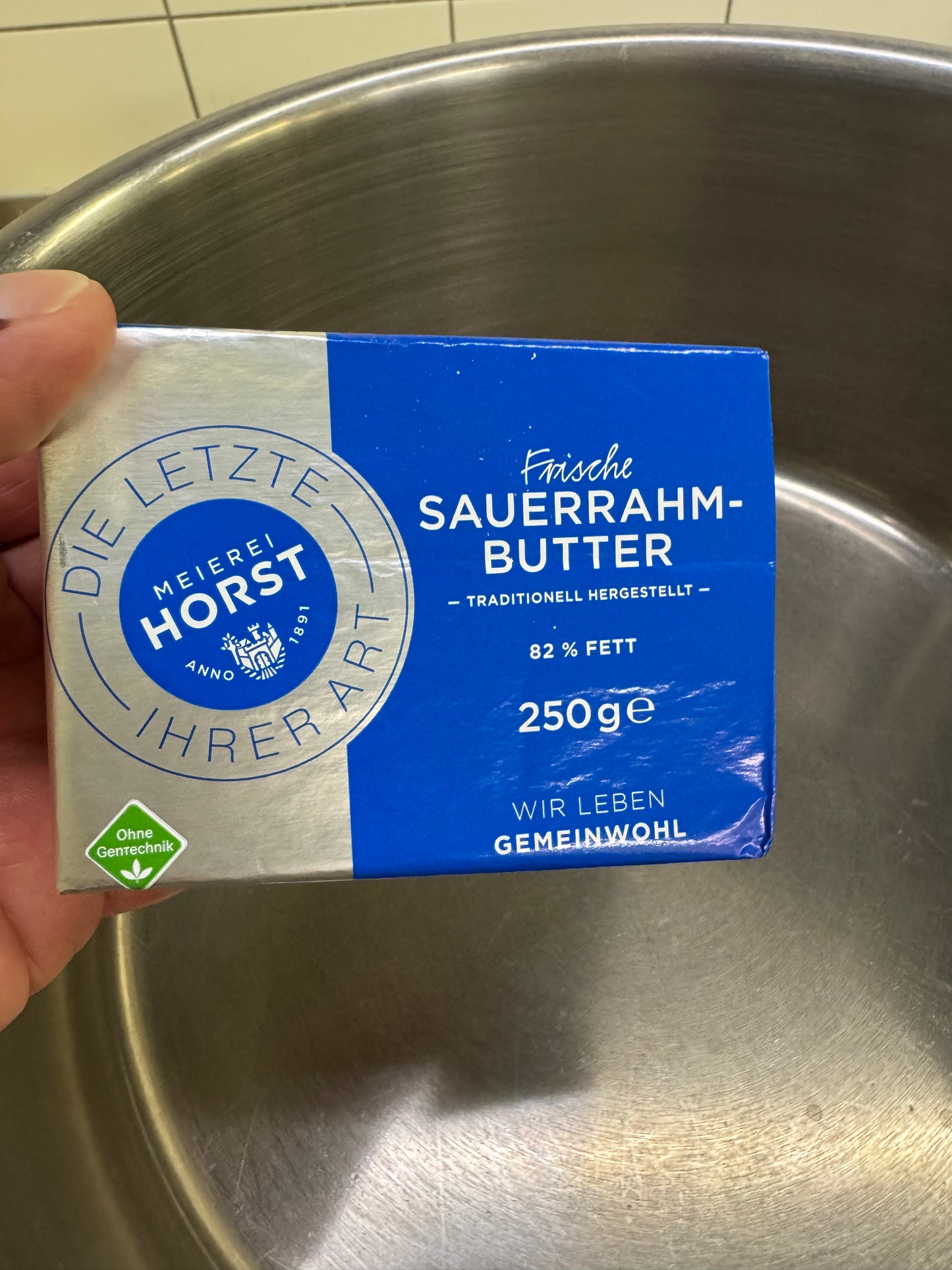 Eine Hand hält eine 250g Packung ECHTE SAUERRAHMBUTTER der Meierei Horst über einen Metalltopf.