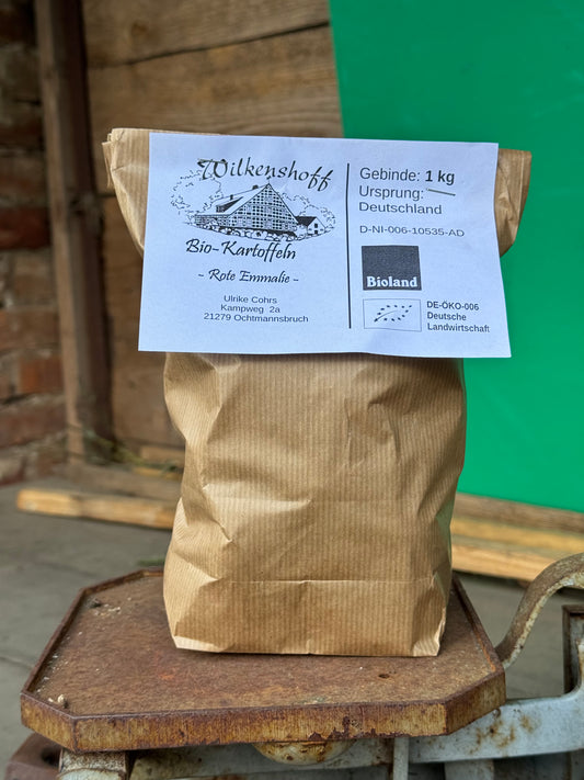 Eine braune Papiertüte mit der Aufschrift „Biohof Wilkenshoff“ enthält eine 1-kg-Charge Kartoffel Rote Emmalie, die elegant auf einer rustikalen Holzoberfläche ruht und den Charme von Sternequalität ausstrahlt.