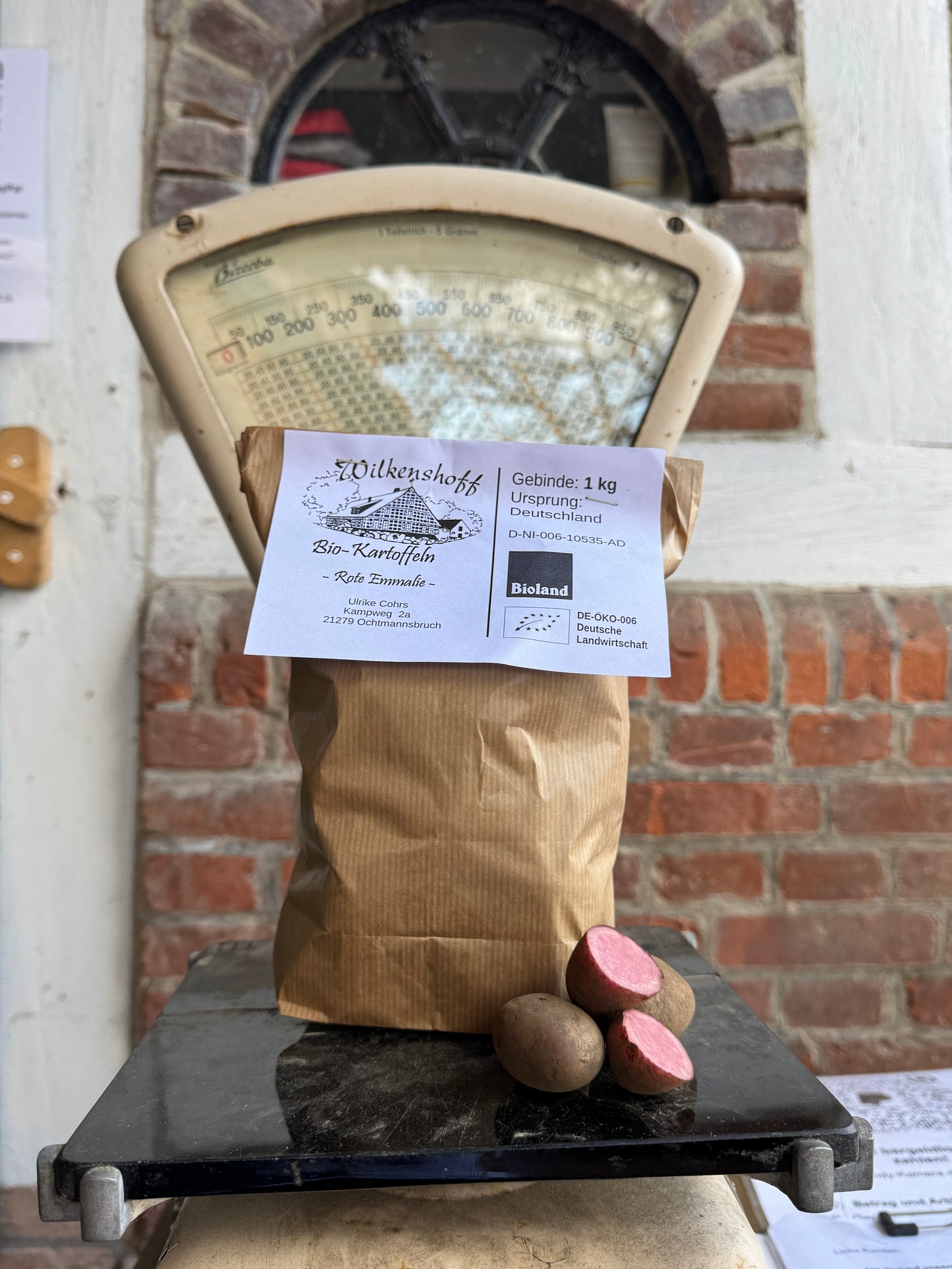 Eine 1-kg-Tüte KARTOFFEL ROTE EMMALIE von Rittmeyers liegt auf einer Vintage-Waage. Zwei ganze und eine halbierte Kartoffel geben den Blick auf das rosafarbene Fruchtfleisch frei und schaffen so eine einladende, rustikale Mahlzeit, die perfekt durch Ihre Lieblingsgewürzmischung ergänzt wird.