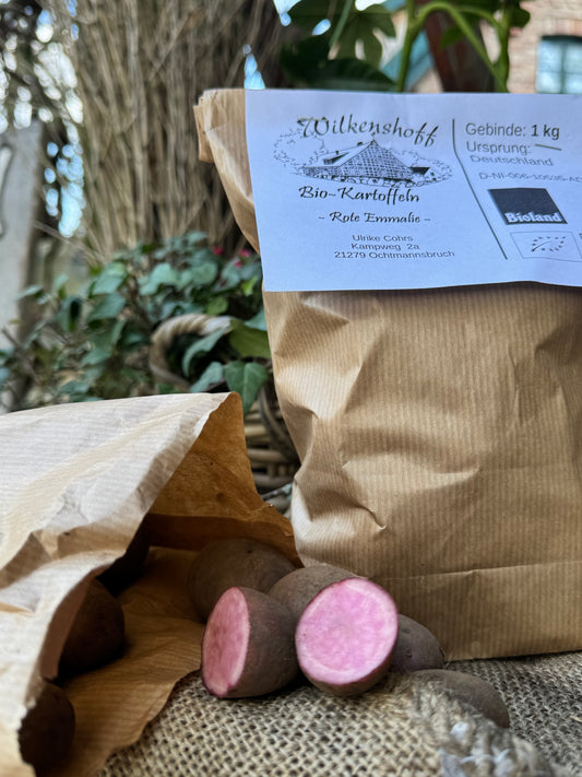 Die KARTOFFEL ROTE EMMALIE - 1 KG braunen Beutel vom Biohof Wilkenshoff mit rosa Kartoffelscheiben auf Jute strahlen Sternequalität aus und sind von leuchtenden Pflanzen umgeben.