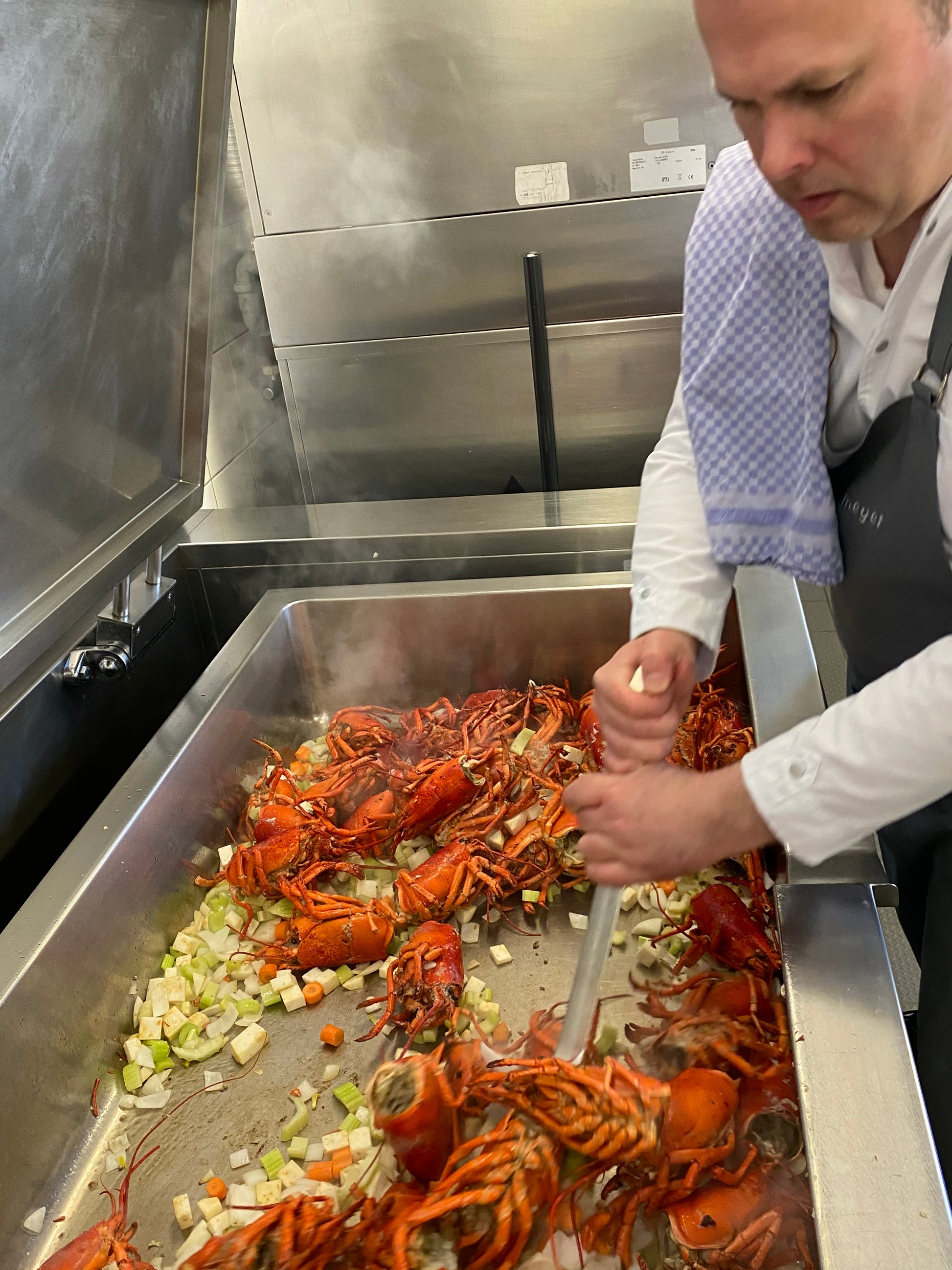 Ein Koch rührt Hummer und gehacktes Gemüse in einer dampfenden Pfanne und bereitet Rittmeyers Besondere Raffinessen HUMMER-ZITRONENGRASSUPPE in einer geschäftigen Großküche zu.