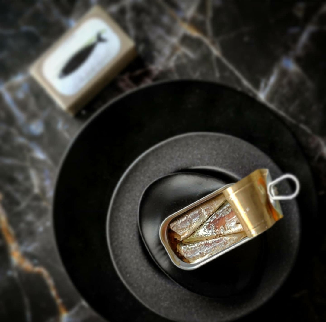 Eine Dose mit „Kleinen Makrelen in Olivenöl“ von José Gourmet steht auf schwarzem Keramikgeschirr neben einer Schachtel auf einer dunklen, strukturierten Oberfläche, und in der Luft liegt der Duft von portugiesischem Olivenöl.