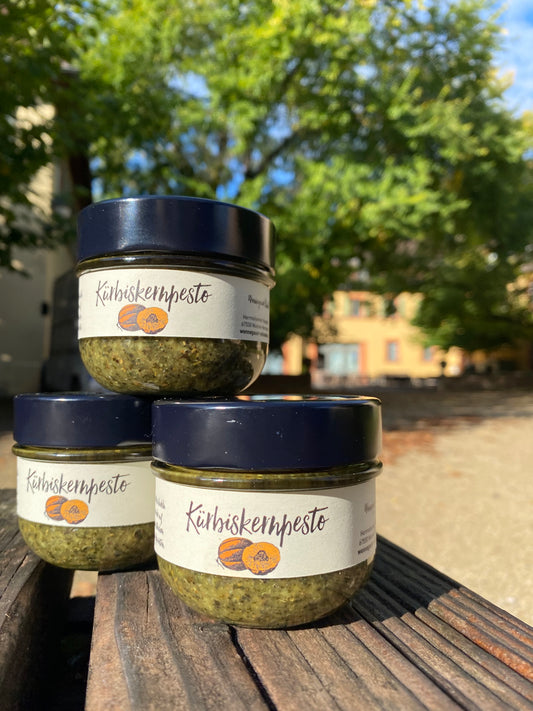 Die KÜRBISKERN-PESTO-Gläser der Wonnegauer Ölmühle sind im Freien auf einer Holzbank gestapelt, vor der ruhigen Kulisse von Bäumen und Gebäuden in der Ferne.