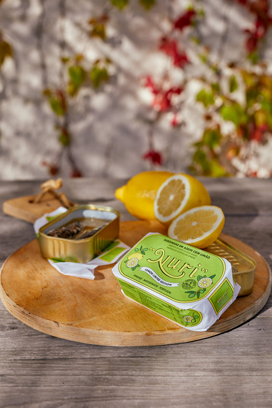 Auf einem Holztisch liegt ein Brett mit NURI SARDINEN MIT OLIVENÖL UND ZITRONE, pikanten Zitronenscheiben und einer rechteckigen, grün umrandeten Verpackung.
