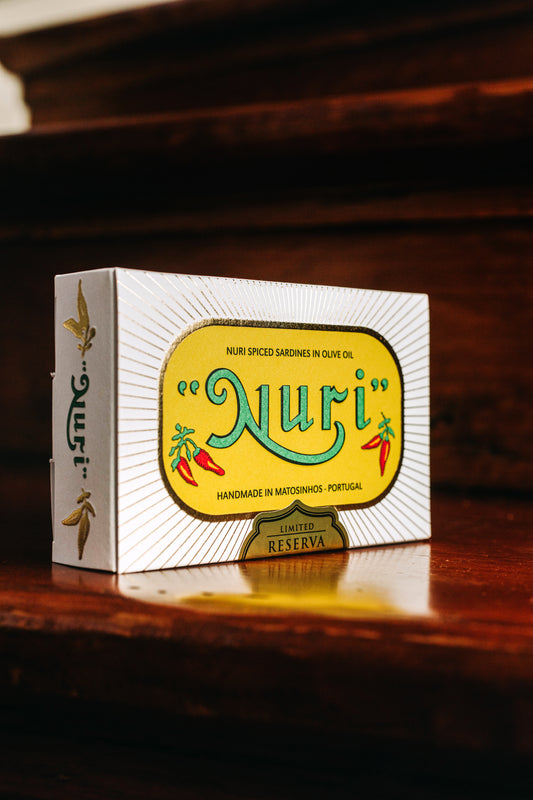 Eine Schachtel NURI PIKANTE JAHRGANGS-SARDINEN RESERVA 2020 in Olivenöl ruht auf einer Holzoberfläche und zeigt lebendige Grafiken und Texte, ein Beispiel für exquisite portugiesische Konservenproduktion.
