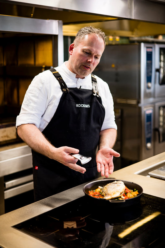 In einer geschäftigen Küche würzt der Koch fachmännisch ein Gericht, während er eine schwarze Schürze trägt. Hinter ihm brummen die Öfen leise, während er die 28 cm große KOCKUMS-BRATPFANNE von Kockums Jernverk benutzt. Der ergonomische Griff aus schwedischem Kohlenstoffstahl sorgt für Präzision und Komfort.