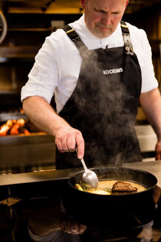 In weißem Hemd und schwarzer Schürze kocht der Koch konzentriert mit einer Kockums Jernverk BRATPFANNE auf dem Herd. Der ergonomische Griff bietet Komfort und Kontrolle bei der fachmännischen Handhabung des hochwertigen Kohlenstoffstahl-Utensils.