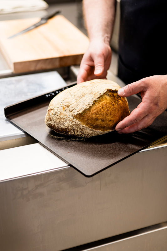 Eine Person hält ein frisch gebackenes Brot auf einer PIZZA/BROT-BACKPLATTE KOCKUMS 37 x 32 cm von Kockums Jernverk in der Küche und betont damit hervorragende Backergebnisse.