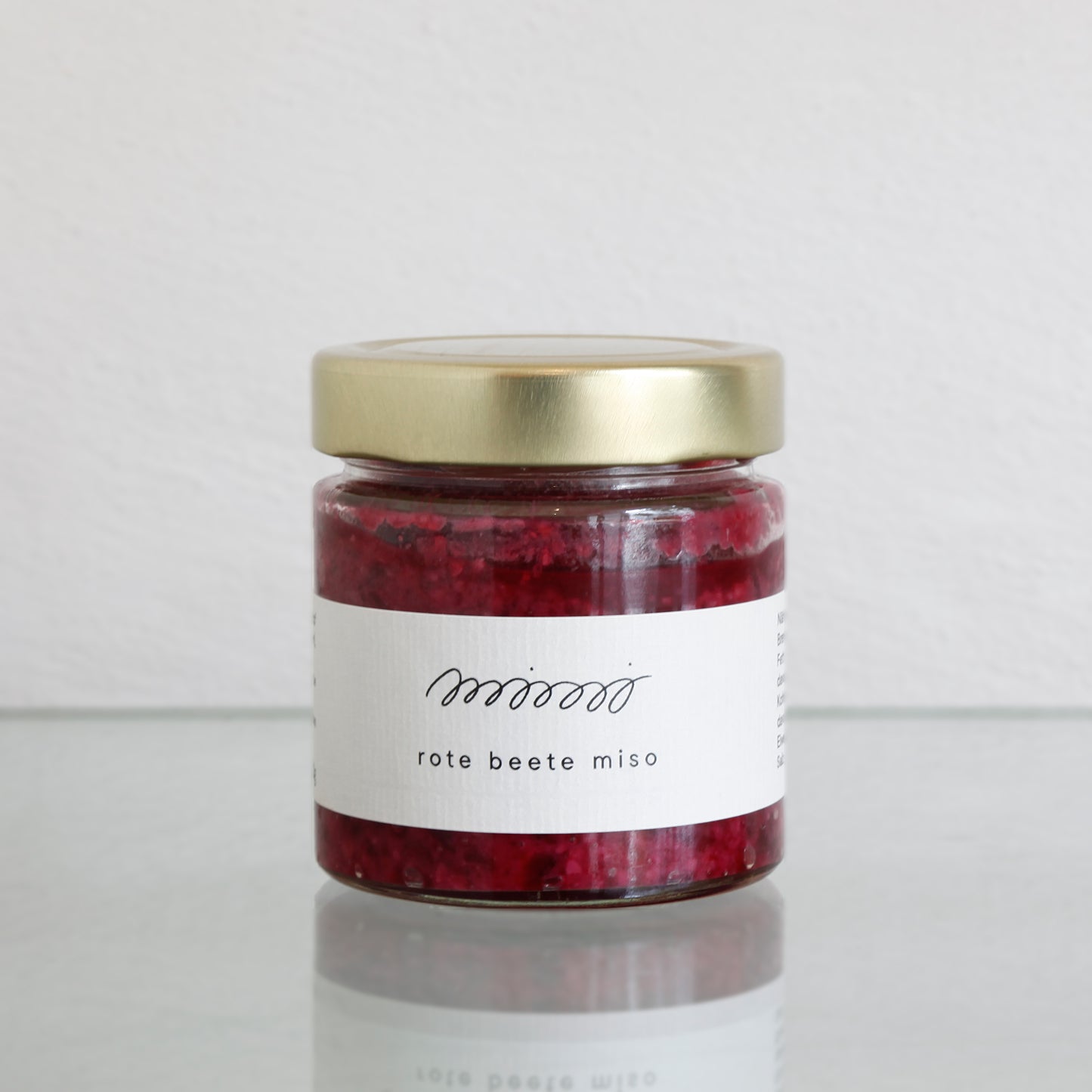 Ein Glas mit Mimi Ferment ROTE BETE MISO und goldenem Deckel steht auf einer Glasoberfläche und verströmt vor einem hellen Hintergrund reiche Umami-Aromen.
