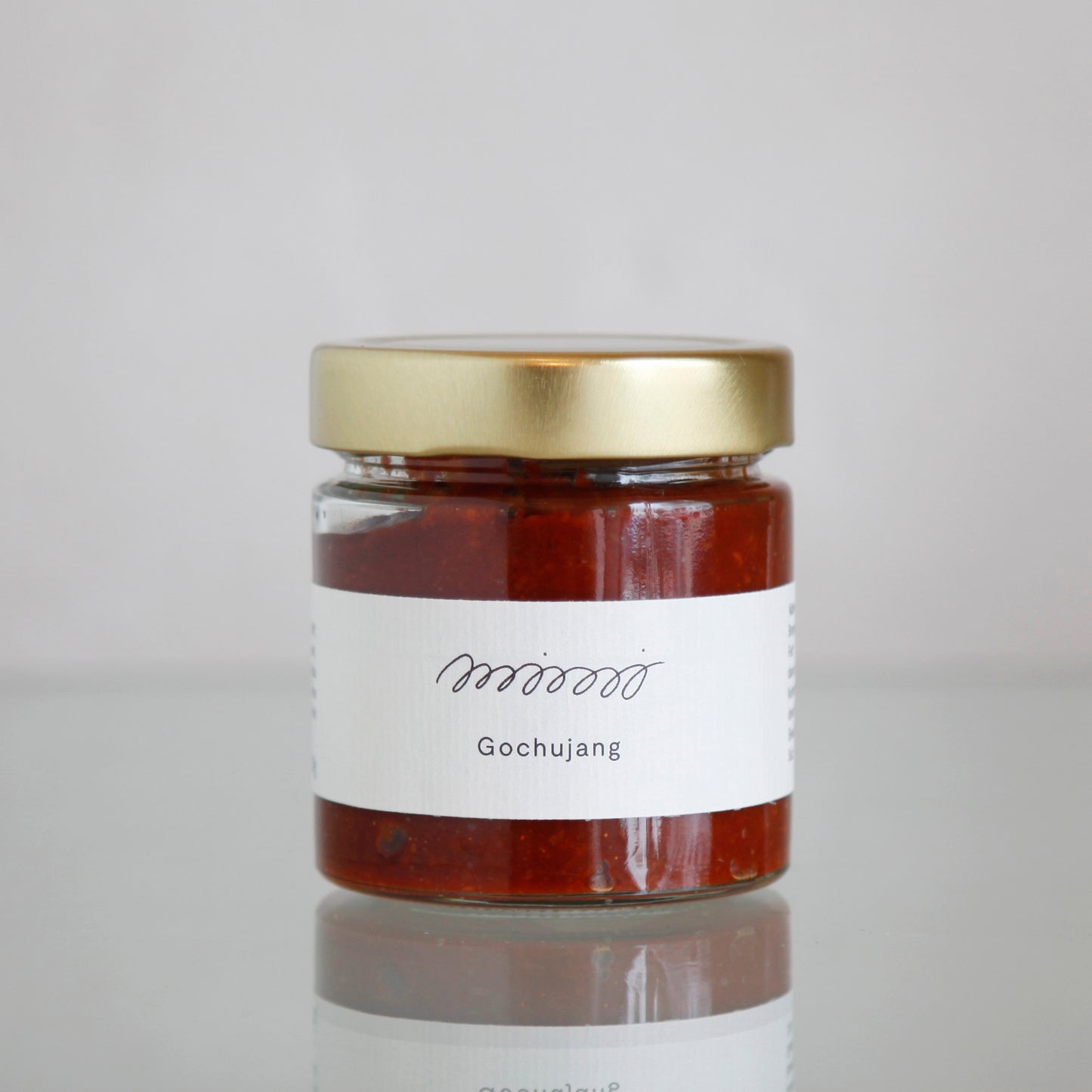 Die GOCHUJANG WÜRZPASTE von mimi ferments glänzt auf einer reflektierenden Oberfläche und ist mit ihrem goldenen Deckel und dem weißen Etikett ein Grundnahrungsmittel der koreanischen Küche, das die reichen Aromen fermentierter Chilipaste zur Geltung bringt.