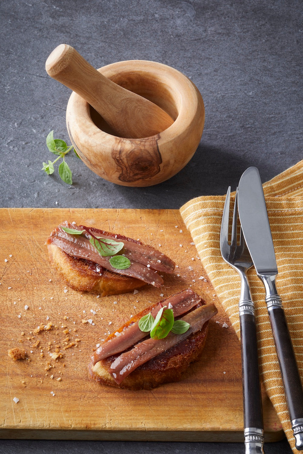 Zwei Bruschetta, belegt mit CODESAs handfiletierter Sardelle und duftendem Basilikum, liegen auf einem Holzbrett, neben Mörser und Stößel, Messer und Gabel.