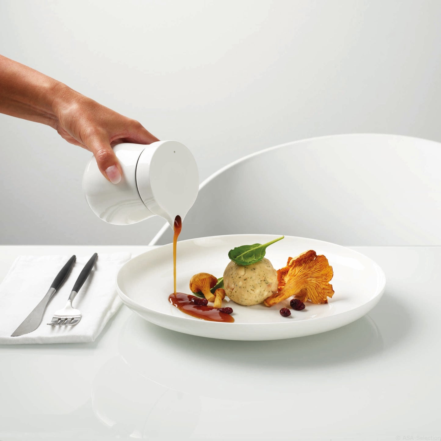 Eine Hand gießt elegant Sauce aus der ASA THERMO SAUCIERE „A TABLE“ von ASA über ein raffiniertes Gericht mit Knödel, Pilzen und Gemüse, wunderschön angerichtet auf feinem Bone China.