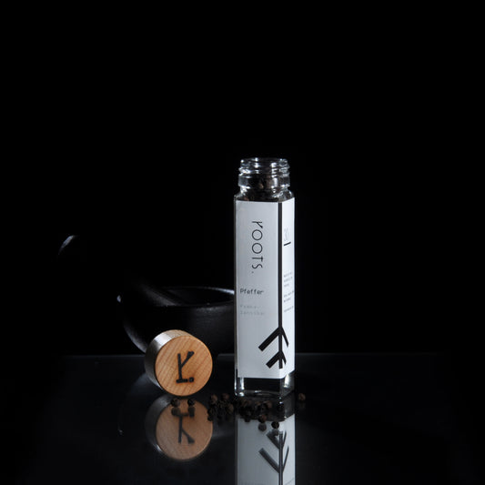 Ein SCHWARZER PEMBA-PFEFFER-Glas von Roots Natural mit Holzdeckel steht inmitten von Pfefferkörnern auf einer reflektierenden Oberfläche und erinnert an den exotischen Reiz von Pemba Sansibar.