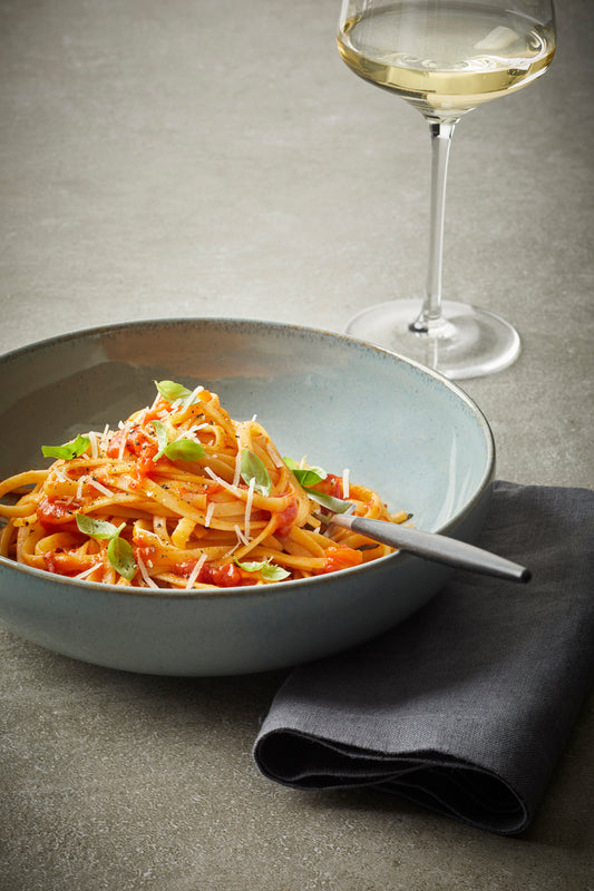 Eine Schüssel Pasta mit Rittmeyers TOMATE FRITO IN OLIVENÖL, Basilikum und Käse, beträufelt mit nativem Olivenöl extra, steht neben einem Glas Weißwein auf einer grauen Serviette.