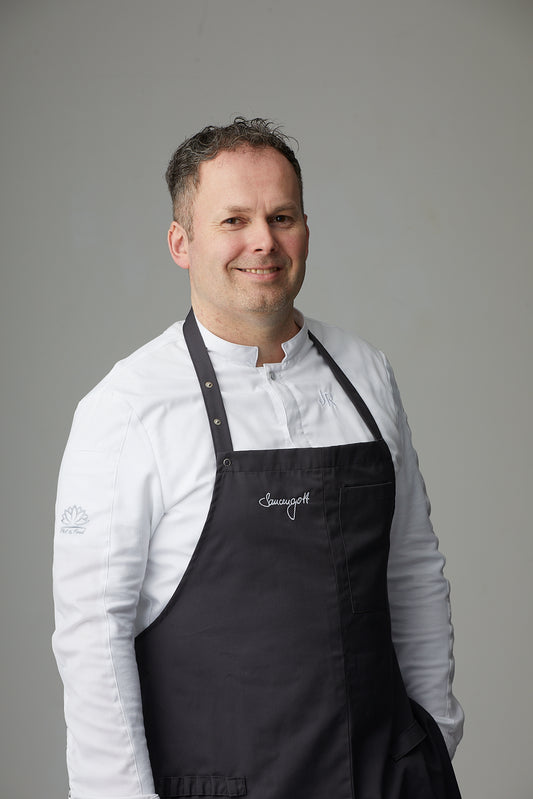 Ein lächelnder „Saucengott“ in Rittmeyers KOCHSCHÜRZE steht vor schlichtem Hintergrund und strahlt kulinarisches Selbstvertrauen aus.