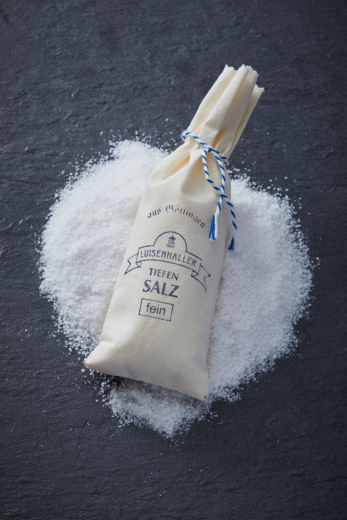 Ein Stoffbeutel mit der Aufschrift „Luisenhaller Tiefensalz“ liegt auf einer dunklen Oberfläche, verstreute Körnchen verraten die Qualität des mineralischen Salzes. Es stammt von Pfannensalinen Europas und verspricht Reinheit und Reichhaltigkeit ohne Zusätze.