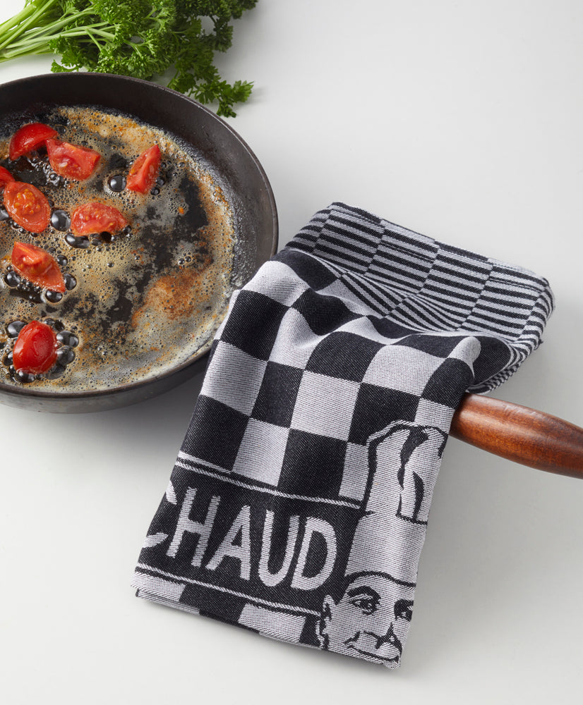 Eine Bratpfanne mit Essen steht neben einem schwarz-weiß karierten Le Chaud devant PREMIUM KÜCHENTUCH - PROFIKÜCHEN-TORCHON, der über den Griff drapiert ist, und frische Petersilie verleiht der Arbeitsplatte einen Hauch von Grün.