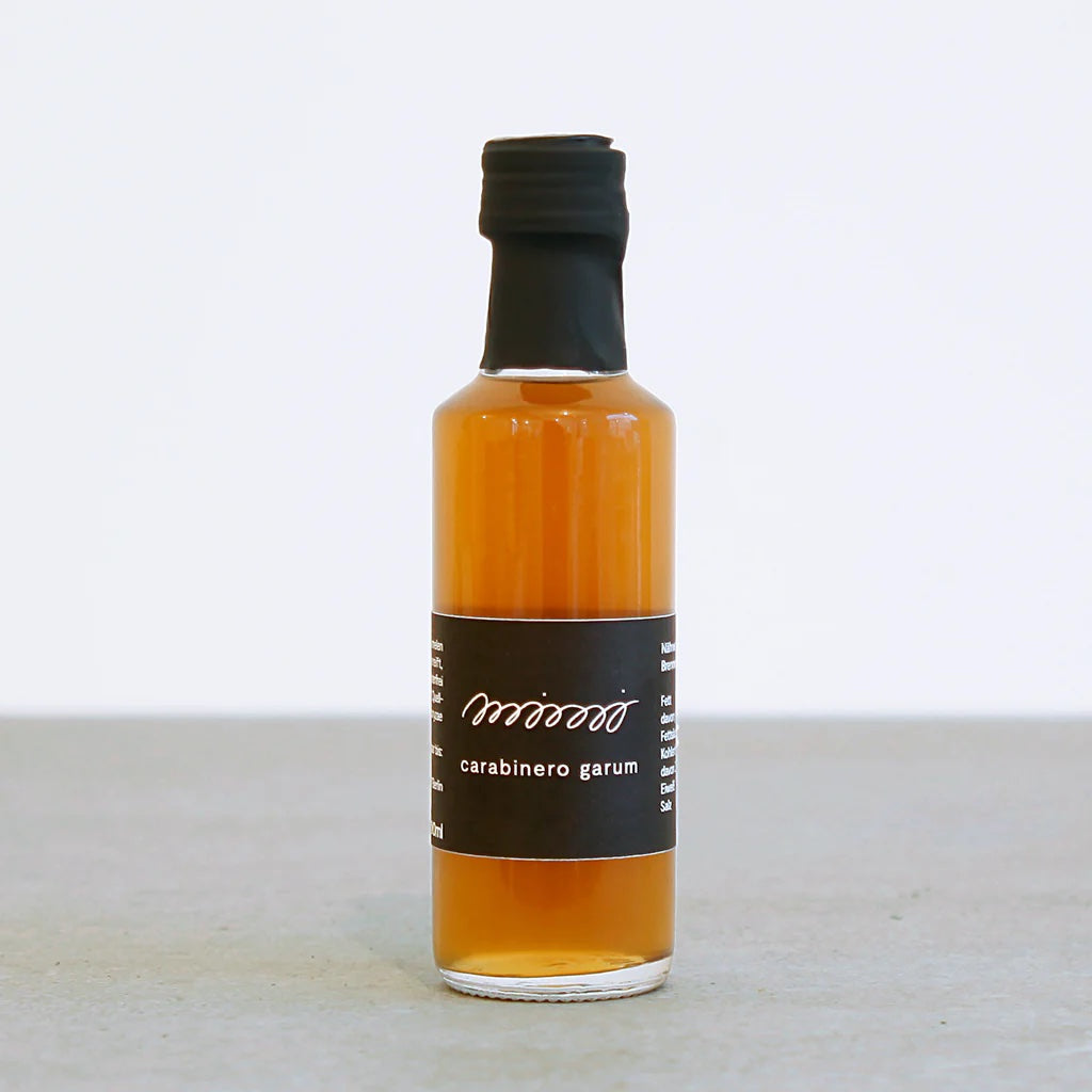 Eine kleine Glasflasche mit bernsteinfarbener Flüssigkeit und einem schwarzen Etikett mit der Aufschrift „mimi ferments CARABINERO LANGOSTINO GARUM – WÜRZSAUCE“, auf dem sich eine fermentierte Würzsauce aus Carabinero-Garnelen befindet.