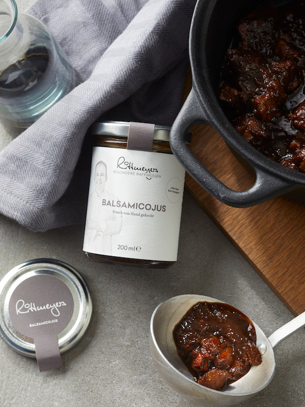 Ein Glas Rittmeyers Besondere Raffinessen BOHNENKRAUTJUS MIT OFENKNOBLAUCH, eine Kelle dunkle Sauce und gegartes Fleisch auf einem Holzbrett – so kommen aromatische Grundbrühen optimal zur Geltung.