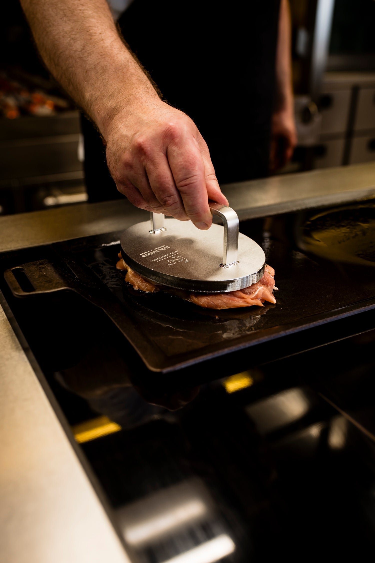 Eine Person verwendet das EDELSTAHL-GRILLGEWICHT von Kockums Jernverk fachmännisch, um beim Grillen von Fleisch in einer Küche knuspriges Braten zuzubereiten.