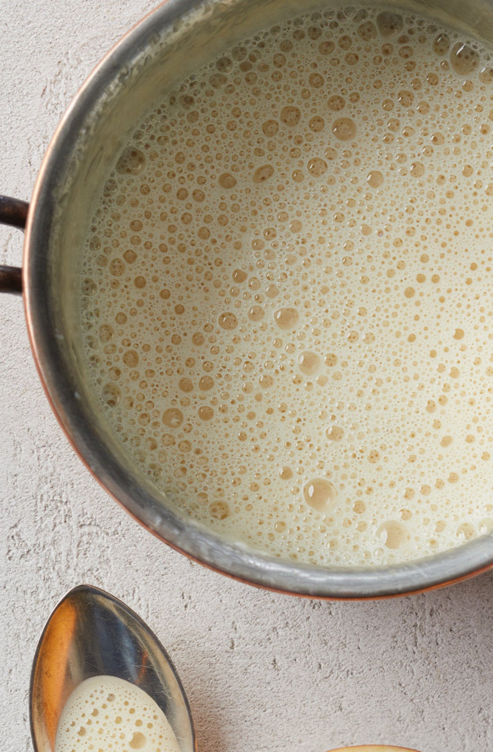 Ein Topf mit schaumiger, cremiger LIMETTEN-INGWERSAUCE Sonderedition von Rittmeyers Besondere Raffinessen steht neben zwei Metalllöffeln auf einer strukturierten Oberfläche.