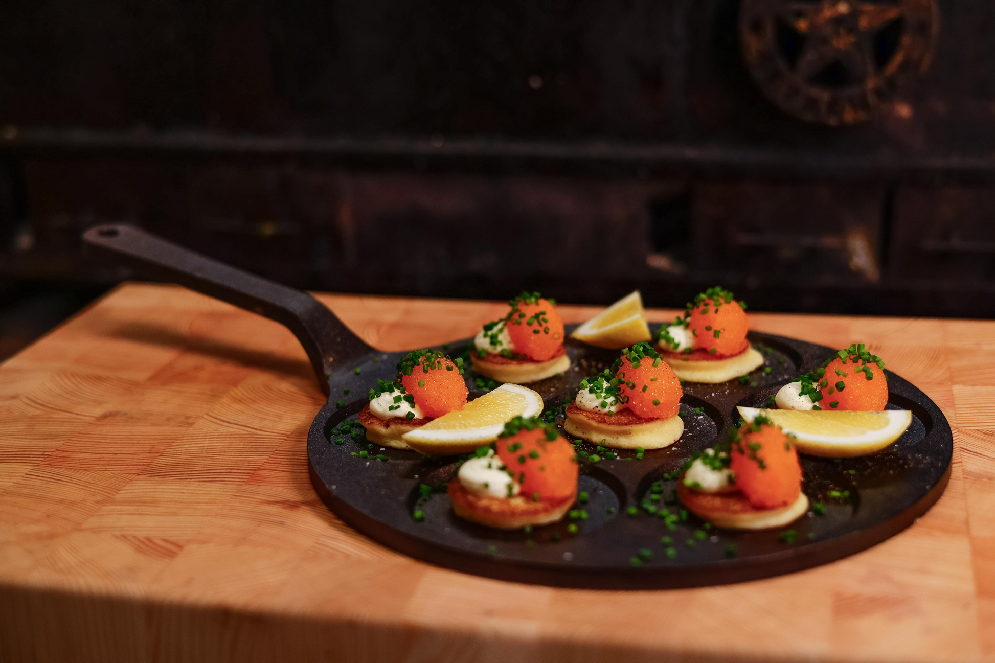 Kleine Blinis mit Sahne, Rogen, Schnittlauch und Zitronenspalten werden auf der Kockums Jernverk MINI-PANCAKE PFANNE 24 cm auf einem Holzbrett serviert.