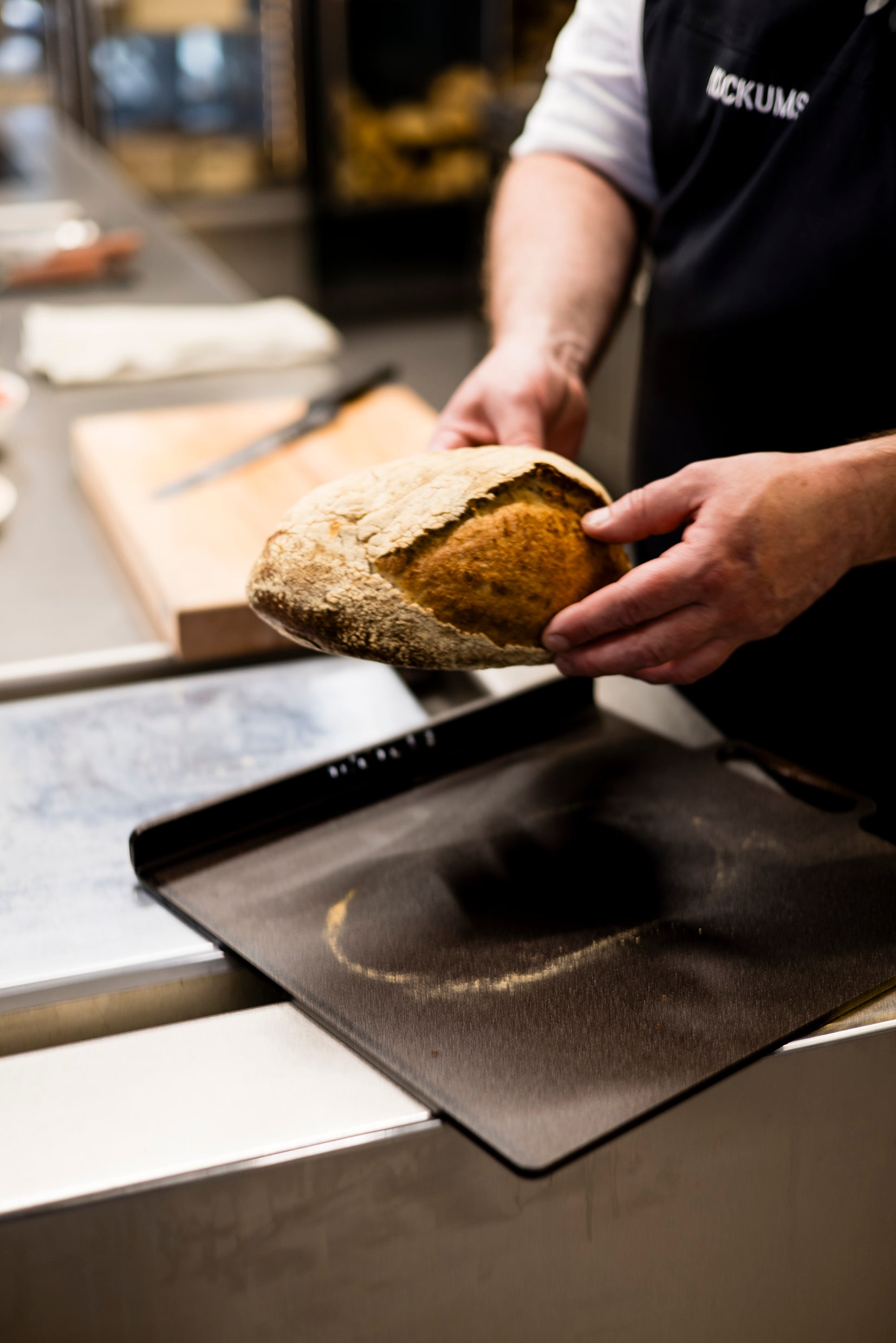 In einer gemütlichen Küche hält jemand rustikales Brot in der Hand, während die Kockums Jernverk PIZZA/BROT-BACKPLATTE 37 x 32 cm daneben ruht.