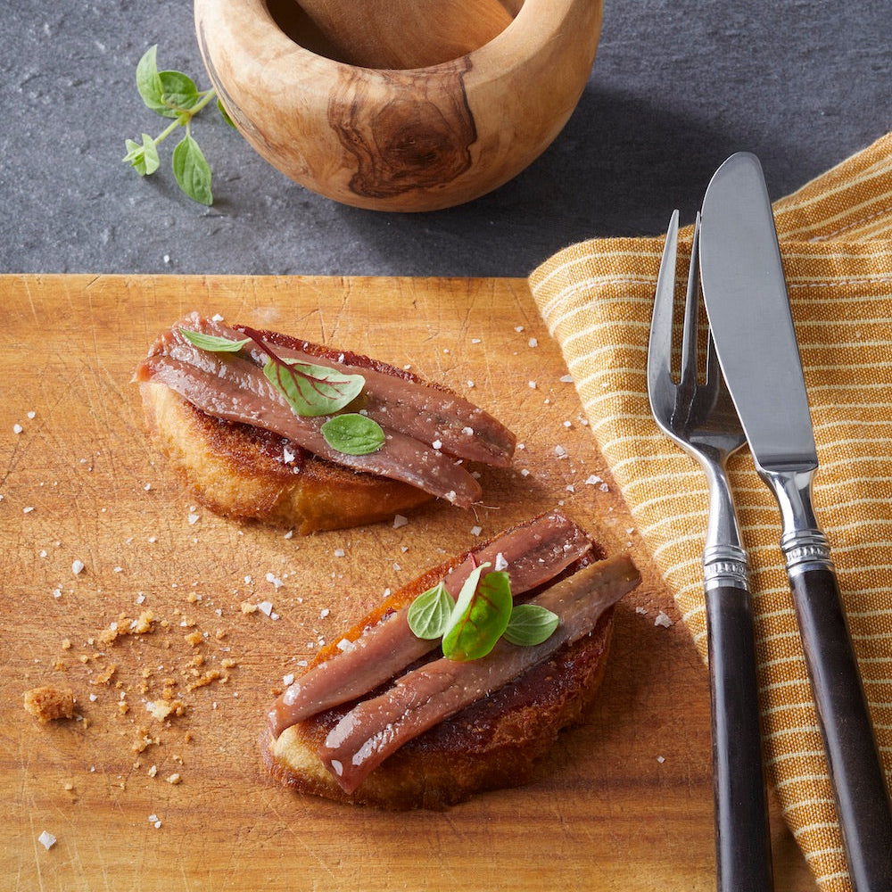 Zwei Bruschetta, belegt mit CODESAs handfiletierter Sardelle und duftendem Basilikum, liegen auf einem Holzbrett, neben Mörser und Stößel, Messer und Gabel.