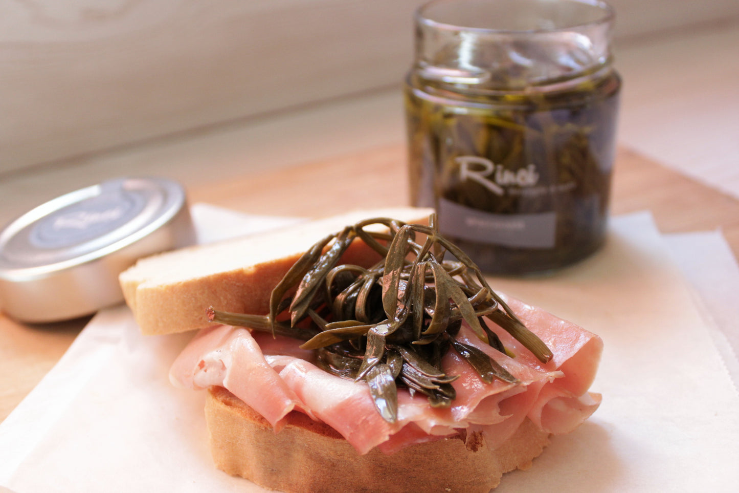Ein Schinkensandwich mit EINGELEGTER MEERFENCHEL liegt neben einem Glas mit Rin's Pickles auf einer Holzoberfläche und ist für zusätzlichen Geschmack mit Olivenöl beträufelt.